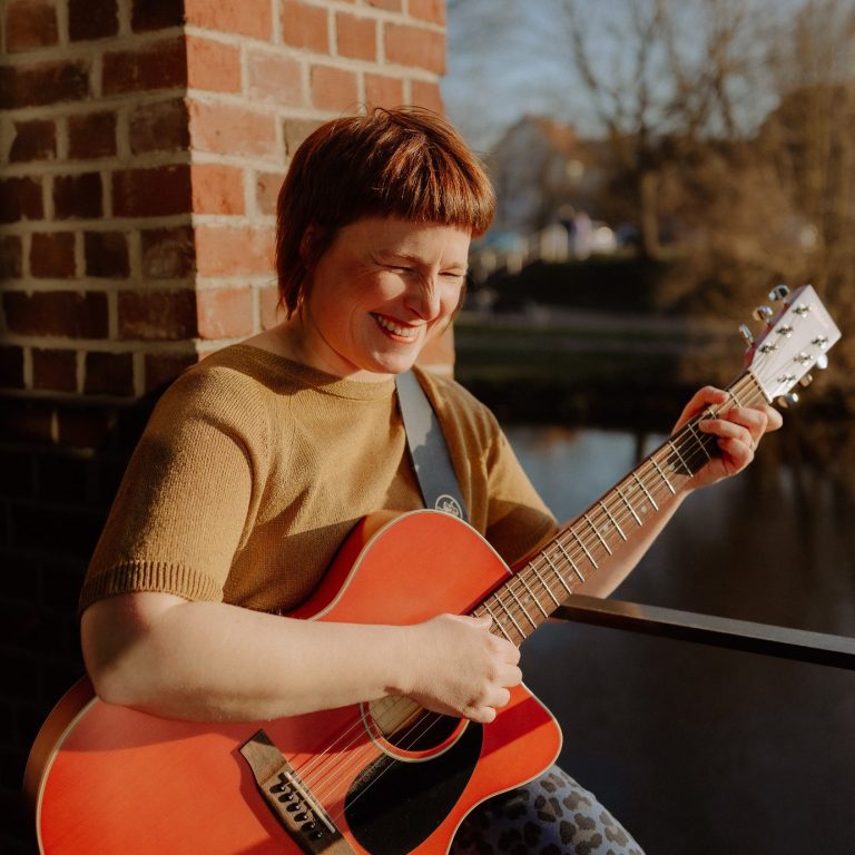 Gesangsunterricht Claudia Neeten spielt draussen Gitarre, im Hintergrund ein Geländer und ein Gewässer