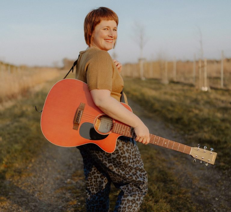 Musik für alle Lebenslagen Claudia Neeten läuft mit ihrer Gitarre umgehängt über einen Feldweg und schaut gut gelaunt zurück in die Kamera.